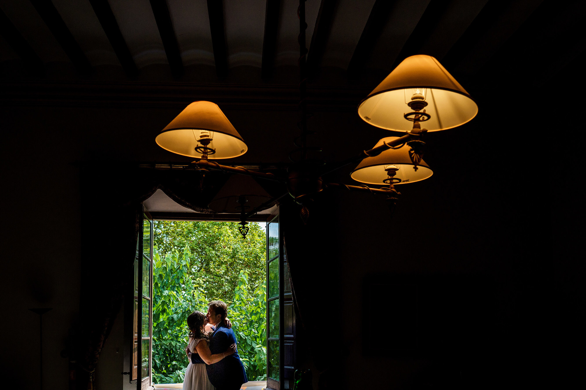 Beso de los novios frente a la ventana de la Masia la Torre del Gall con las luces del techo en primer plano.