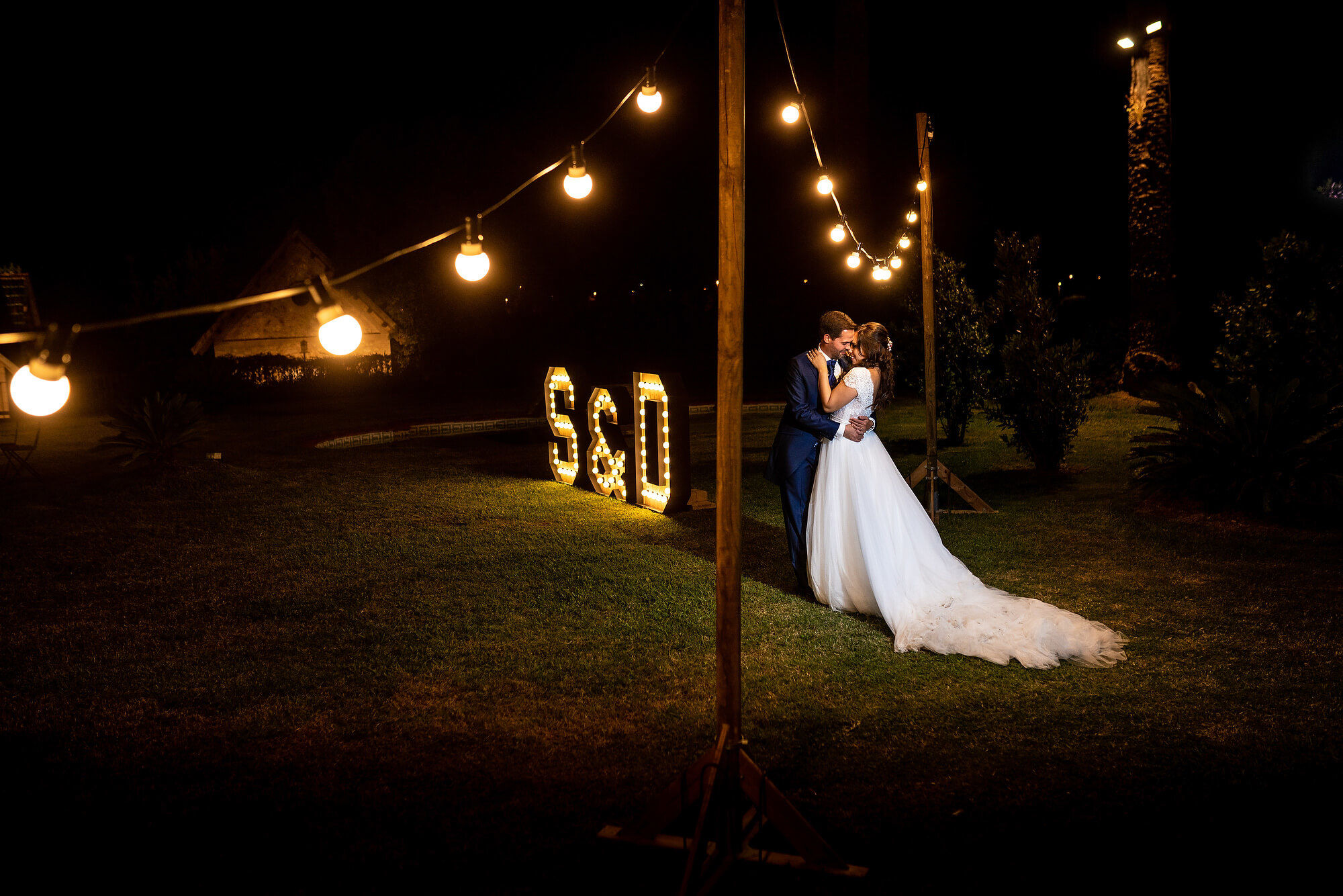 Beso de los novios frente a las letras de madera con bombillas en la boda en Heretat Sabartés.
